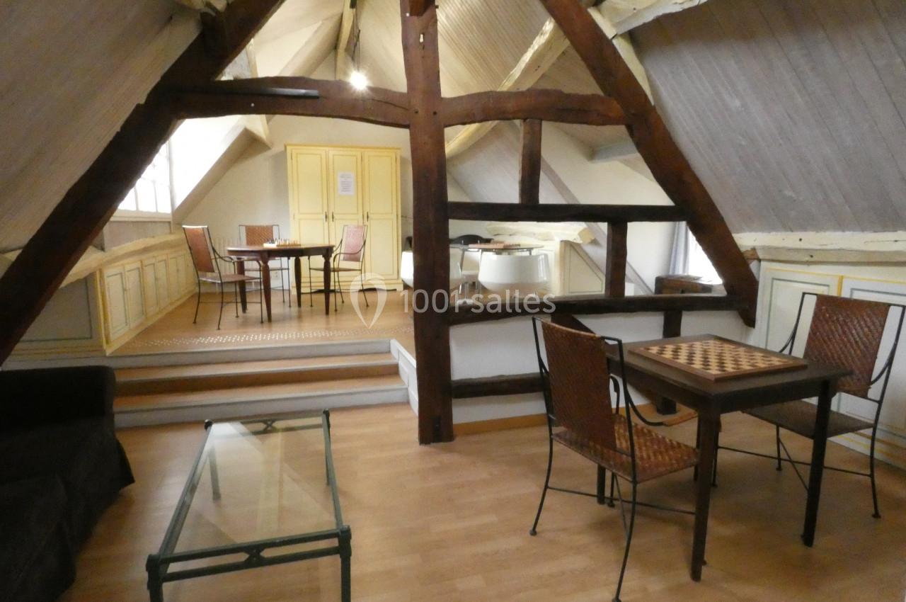 Salle mansardée avec poutres apparentes, mobilier en bois, table de jeu, table à manger et lumière naturelle.