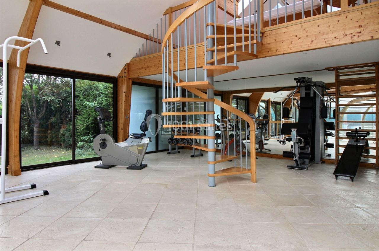 Salle de sport lumineuse avec équipements de fitness, escalier en colimaçon et grandes baies vitrées donnant sur un jardin.