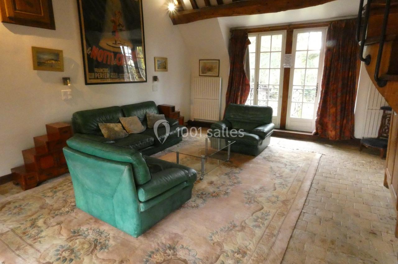 Salon avec canapé et fauteuils verts, table basse en verre, tapis clair et poutres apparentes sous un plafond haut.