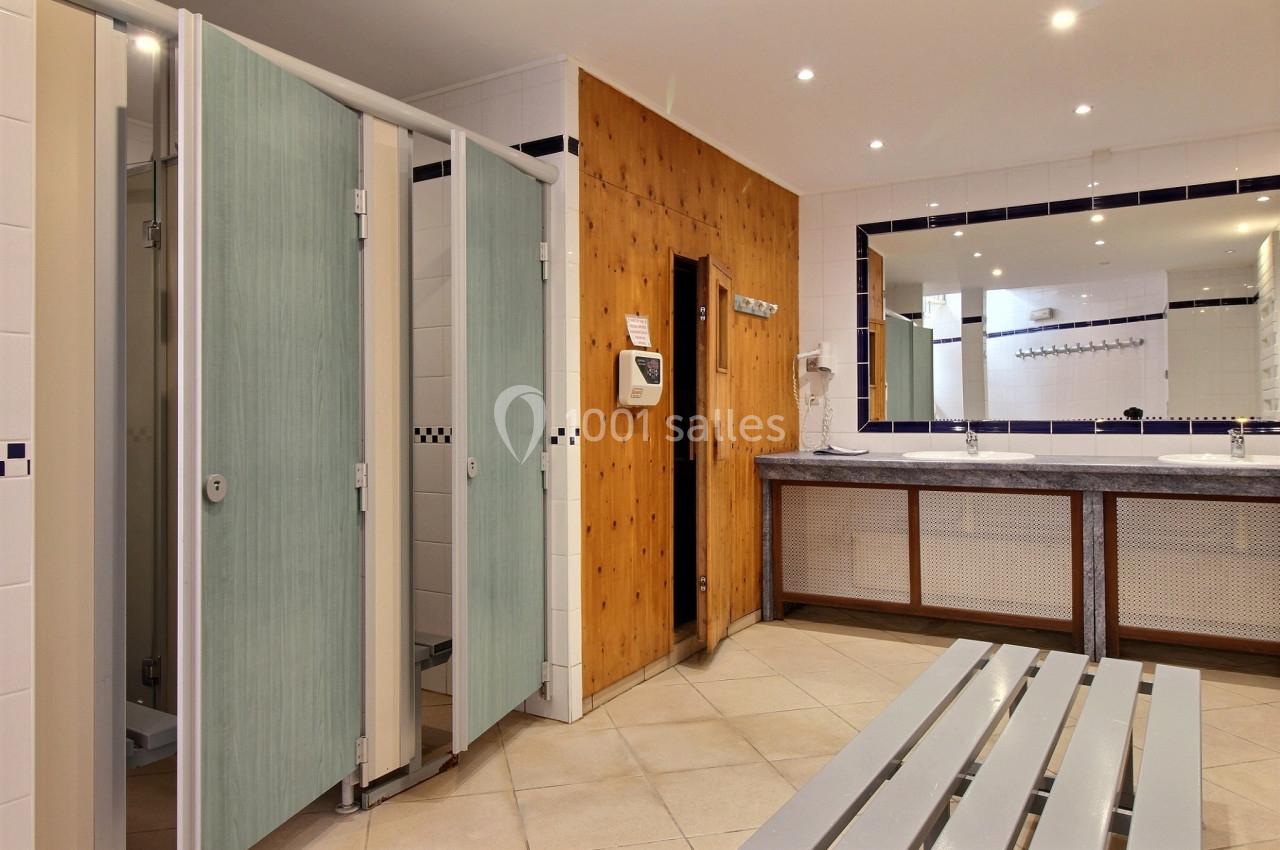 Vestiaires avec cabines de douche, lavabos, miroir mural et banc en métal sur un sol carrelé.