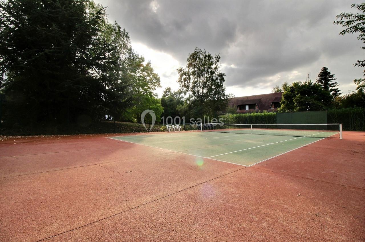 Terrain de tennis extérieur avec revêtement rouge et vert, entouré d'arbres et de végétation sous un ciel nuageux.