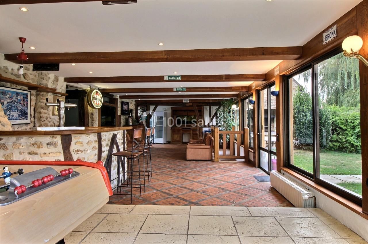 Salle lumineuse avec bar en bois, baby-foot au premier plan, grandes fenêtres donnant sur un jardin.