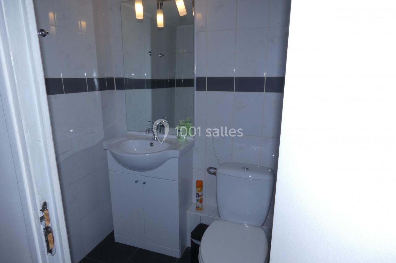 Salle de bain avec lavabo, miroir, éclairage mural, toilettes et carrelage blanc avec bande décorative grise.