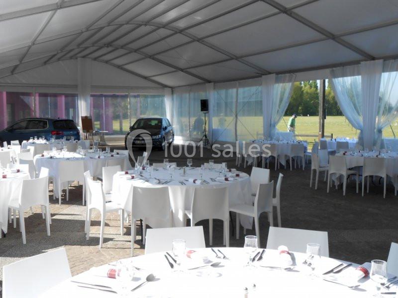 Salle sous chapiteau avec tables rondes dressées pour un événement, voitures garées en arrière-plan.