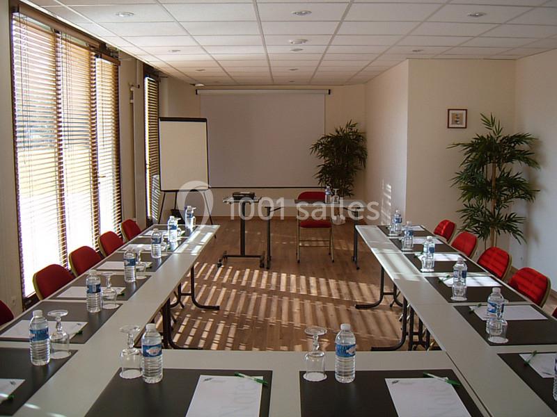 Salle de réunion lumineuse avec tables disposées en U, chaises rouges, écran de projection et fournitures de bureau.