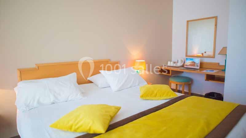 Chambre lumineuse avec un lit double, des coussins jaunes, une coiffeuse en bois et une lampe allumée.