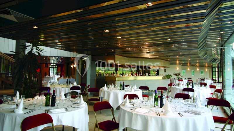 Salle de restaurant élégante avec tables rondes dressées, chaises rouges et un bar en arrière-plan.