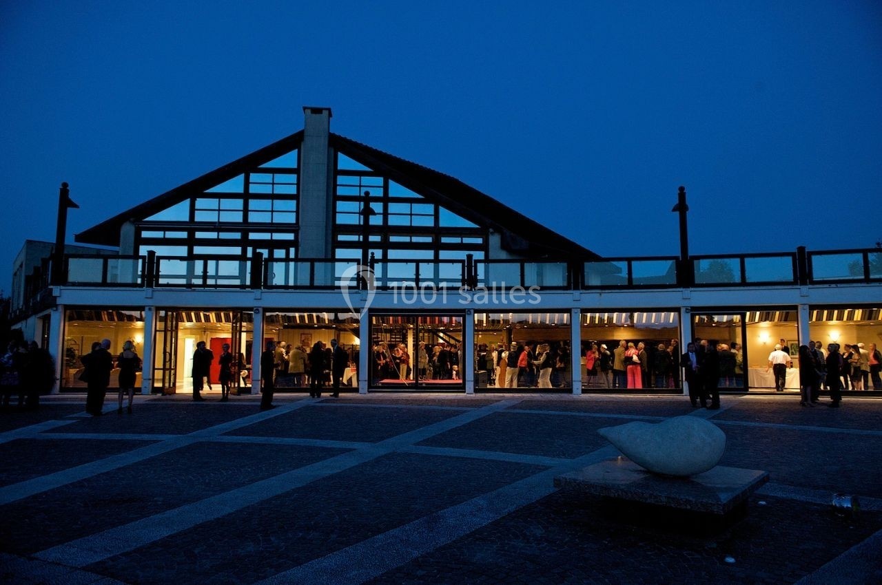 Façade vitrée d'un bâtiment illuminé en soirée, avec des personnes rassemblées à l'intérieur et à l'extérieur.