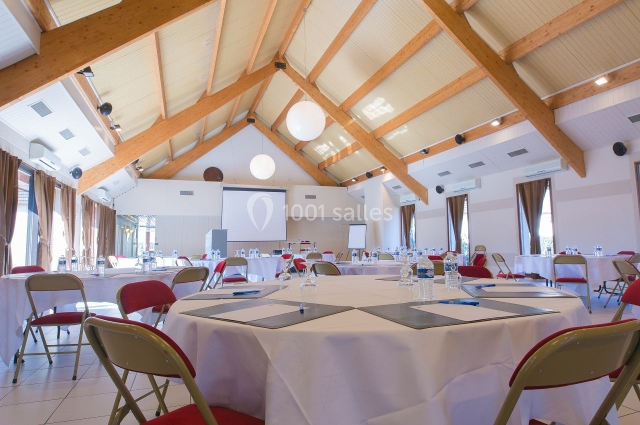 Salle de réunion lumineuse avec tables rondes dressées, chaises pliantes et projecteur installé devant un écran blanc.