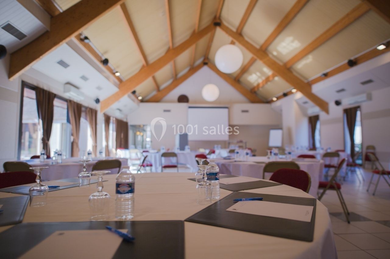 Salle de réunion lumineuse avec tables rondes dressées, chaises, écran de projection et grandes fenêtres.