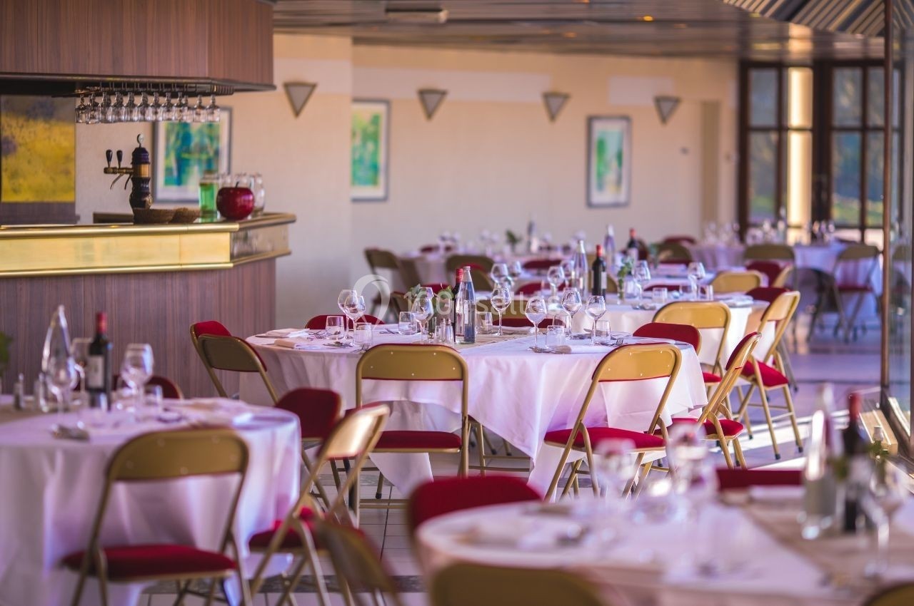 Salle de restaurant lumineuse avec tables dressées, nappes blanches, chaises rouges et un bar en arrière-plan.