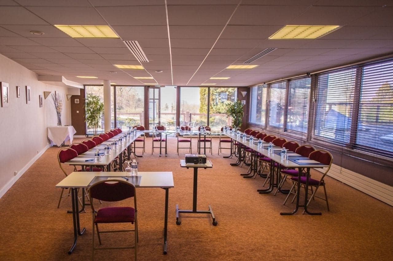 Salle de réunion lumineuse avec tables disposées en U, chaises rouges, projecteur et vue sur l'extérieur.