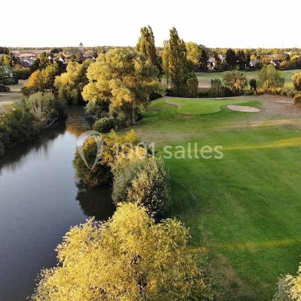 Vue aérienne d'un parcours de golf entouré d'arbres et d'un plan d'eau, avec des habitations en arrière-plan.