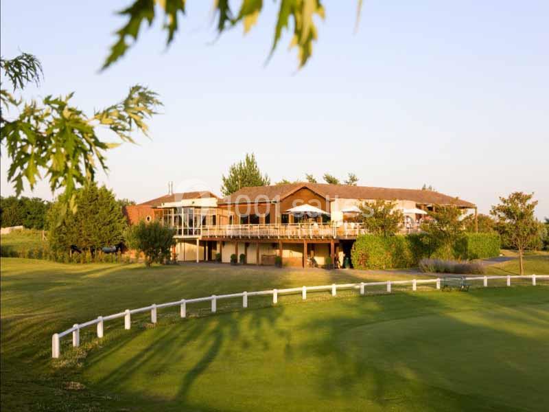 Bâtiment en bois avec terrasse surplombant un terrain verdoyant, entouré d'arbres sous un ciel dégagé.