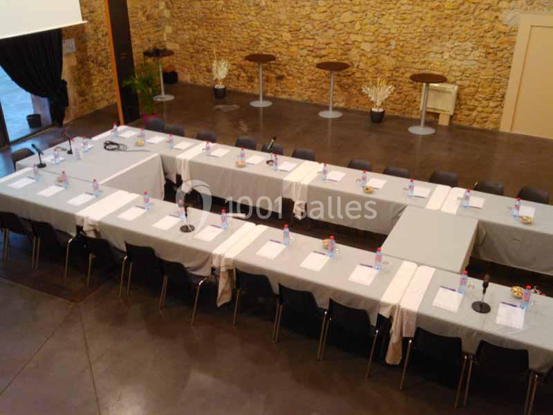 Disposition en U de tables couvertes de nappes blanches, équipées de micros, bouteilles d'eau et documents, dans une salle…