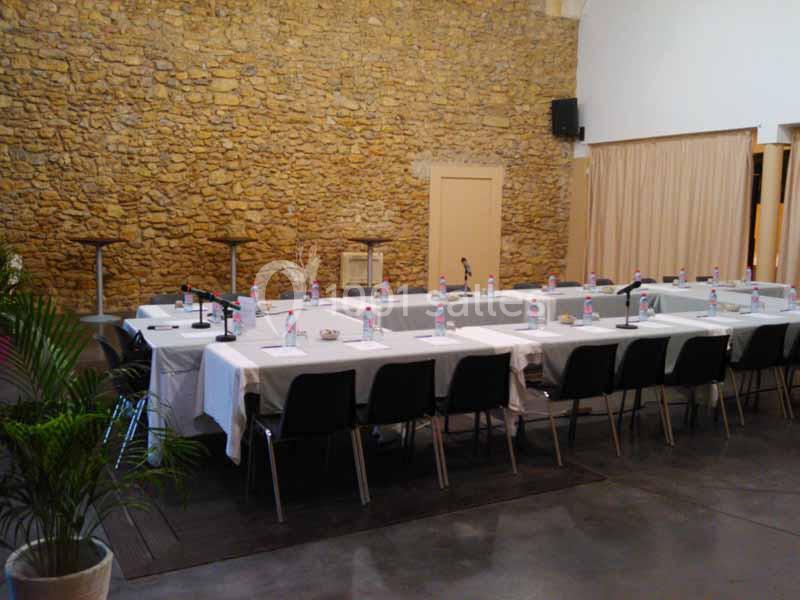 Salle de réunion avec des tables disposées en U, équipées de microphones et de bouteilles d'eau, devant un mur en pierre.