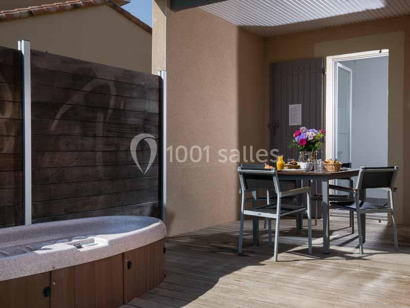 Terrasse en bois avec table dressée, chaises, bain à remous et porte ouverte sur une pièce lumineuse.