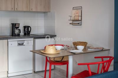 Salle à manger lumineuse avec table en bois, chaises colorées, cuisine ouverte et décoration sobre.