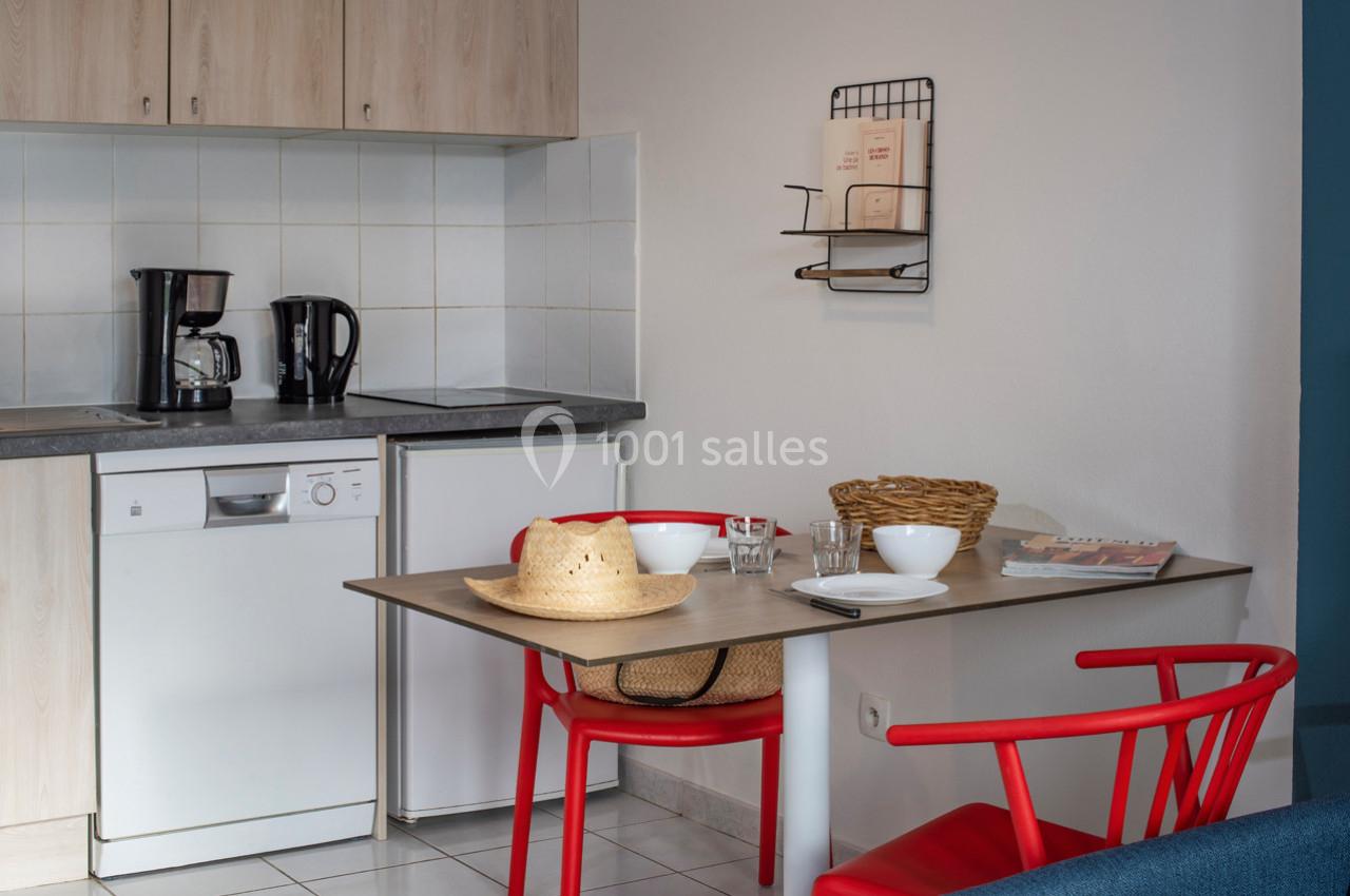 Cuisine avec table en bois, deux chaises rouges, vaisselle, cafetières, et étagère murale avec livres.