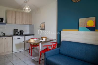 Salle à manger lumineuse avec table en bois, chaises colorées, cuisine ouverte et décoration sobre.