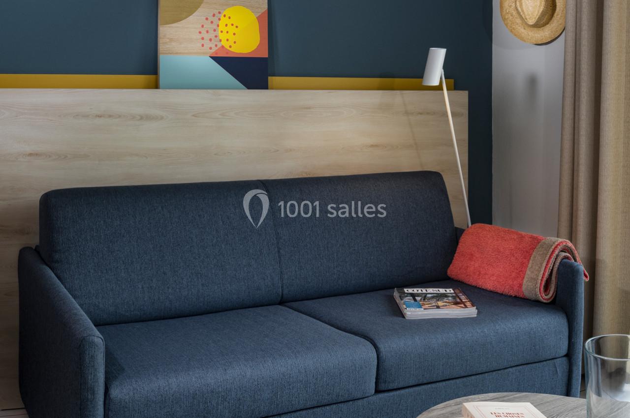 Canapé bleu avec plaid rouge, table basse avec livres et verre, mur bleu décoré d'un tableau et d'un chapeau suspendu.