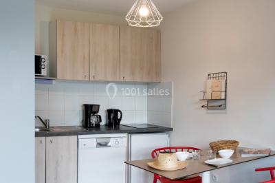 Salle à manger lumineuse avec table en bois, chaises colorées, cuisine ouverte et décoration sobre.