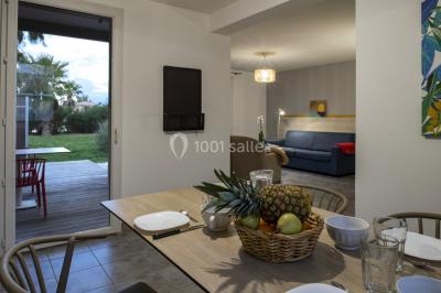 Salle à manger lumineuse avec table en bois, chaises colorées, cuisine ouverte et décoration sobre.