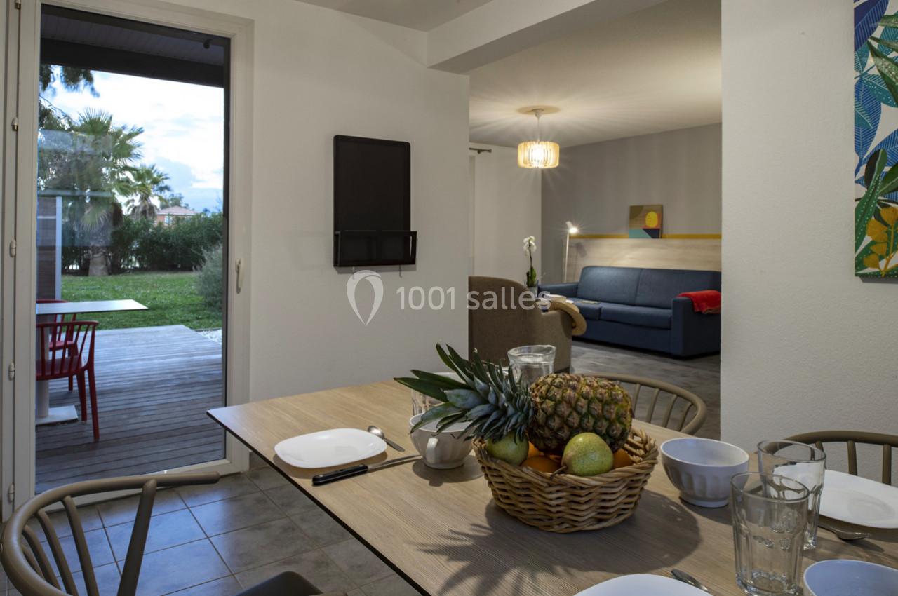 Vue d'une salle à manger avec une table dressée, un salon en arrière-plan et une terrasse donnant sur un jardin.