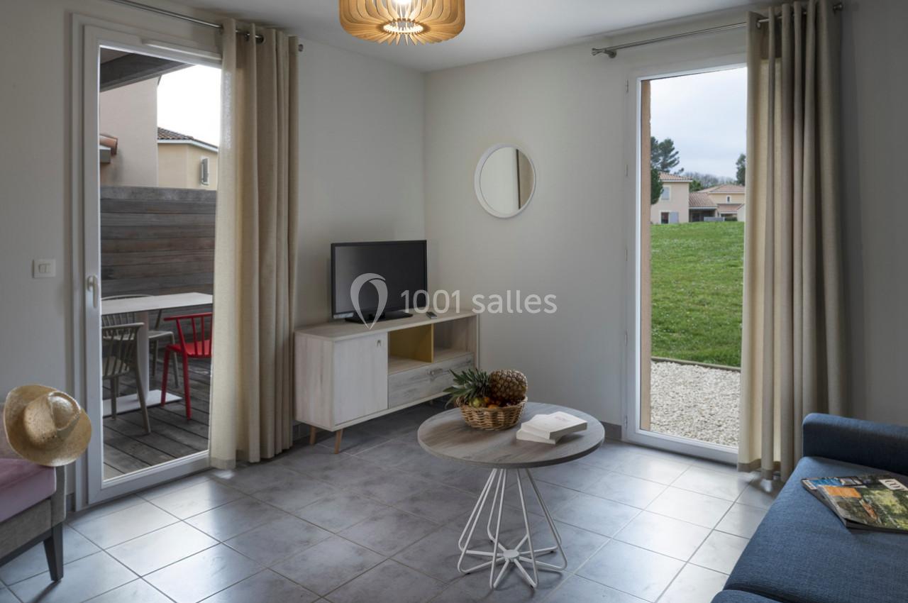 Salon lumineux avec canapé, table basse, télévision, grandes baies vitrées donnant sur une terrasse et un jardin.