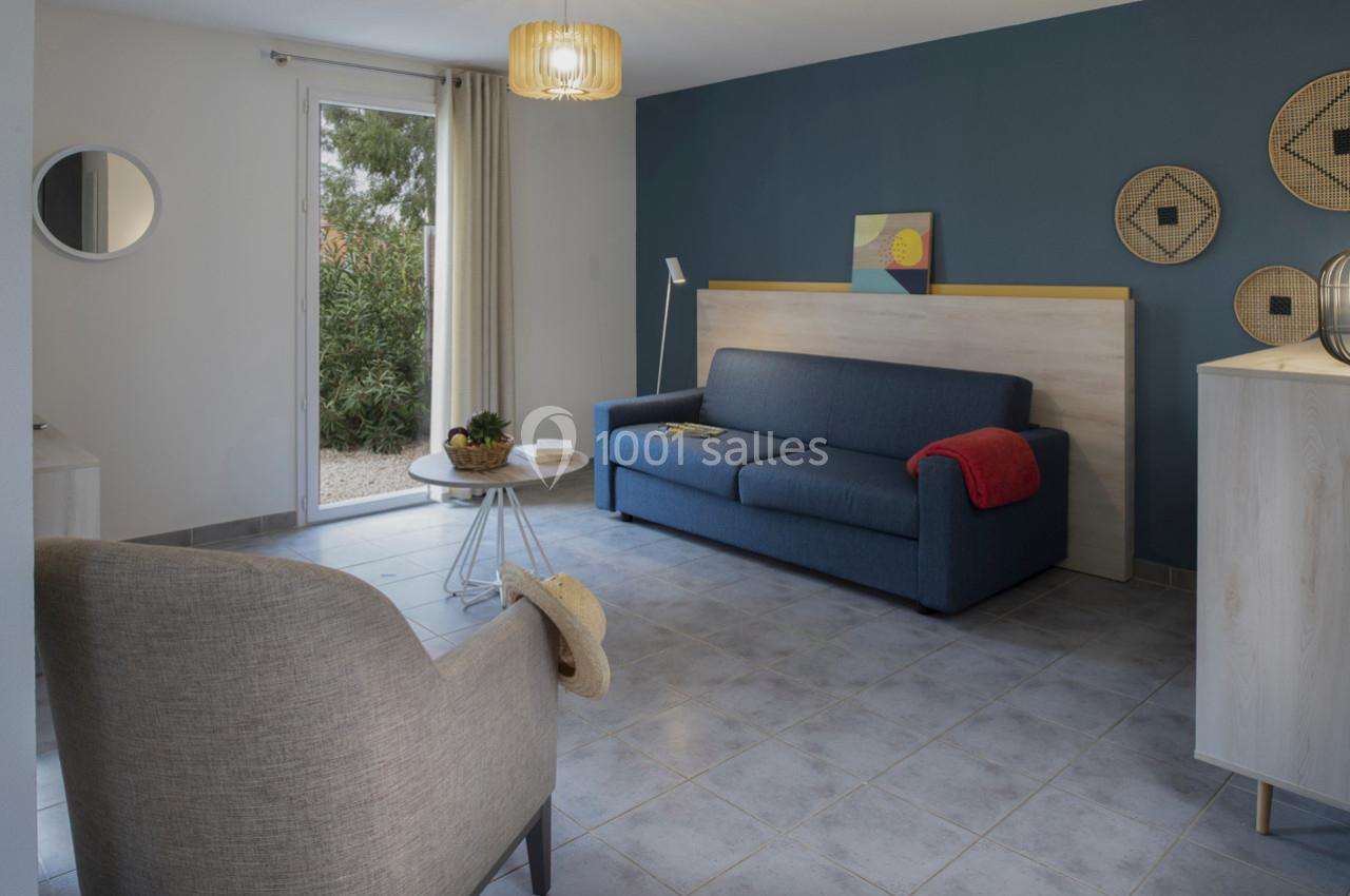 Salon lumineux avec canapé bleu, fauteuil beige, table basse ronde, mur bleu décoré et baie vitrée donnant sur un jardin.