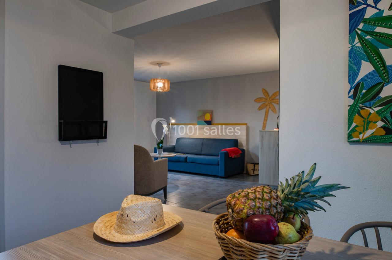 Vue intérieure d'une pièce lumineuse avec un salon en arrière-plan et une table décorée d'un panier de fruits au premier…