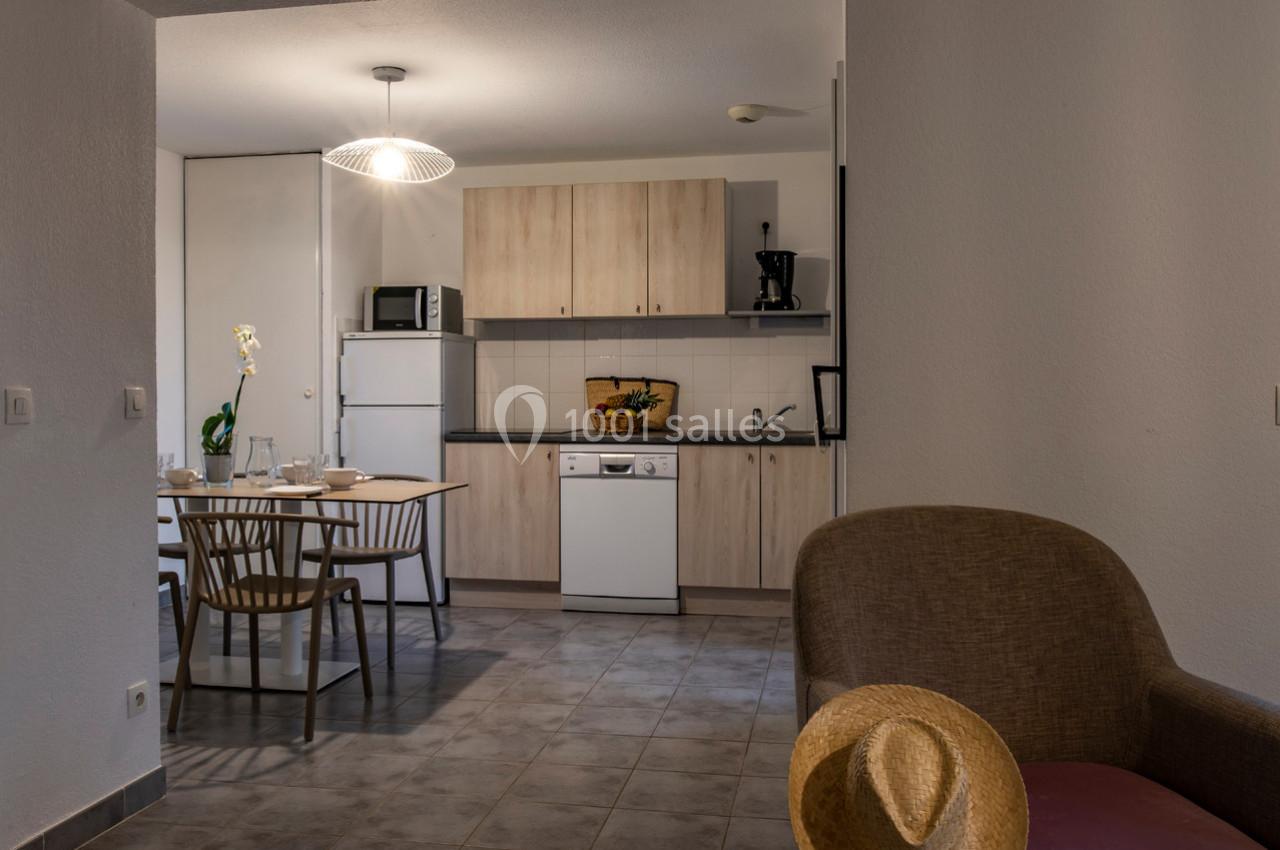 Cuisine ouverte avec mobilier en bois clair, table dressée, électroménagers et fauteuil avec un chapeau posé dessus.