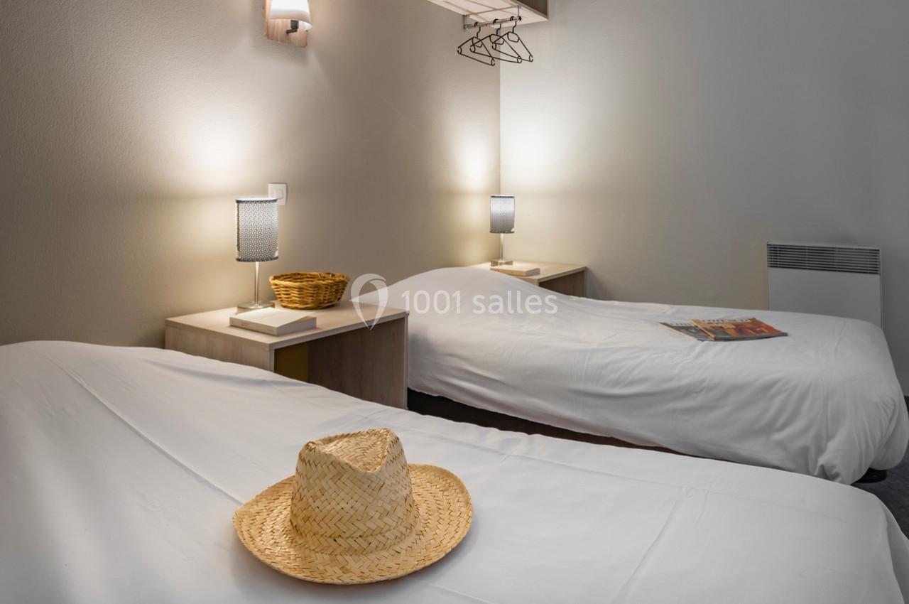Deux lits simples avec draps blancs, chapeaux en paille et lampes de chevet allumées dans une chambre sobre.