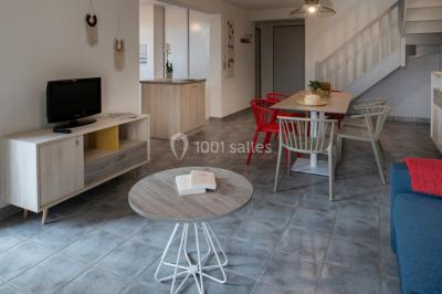 Salle à manger lumineuse avec table en bois, chaises colorées, cuisine ouverte et décoration sobre.