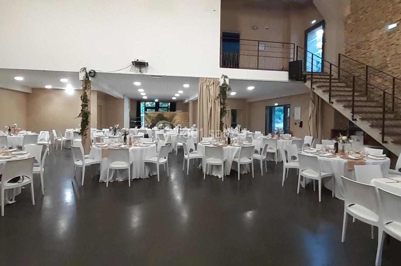 Salle de réception décorée avec des tables rondes et rectangulaires dressées pour un événement, éclairage doux.