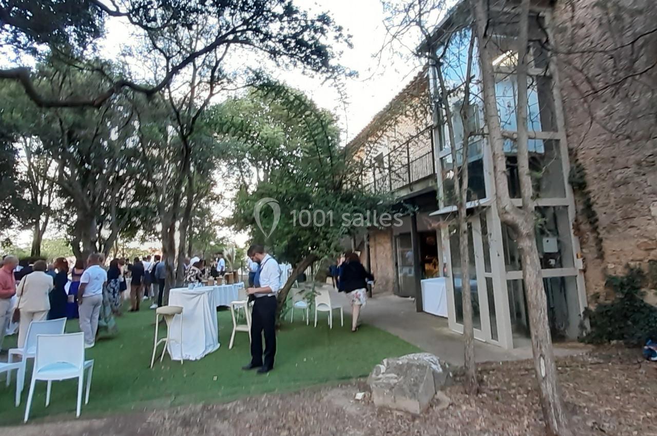 Groupe de personnes rassemblées dans un jardin avec des tables hautes, près d'un bâtiment en pierre et de grandes baies…