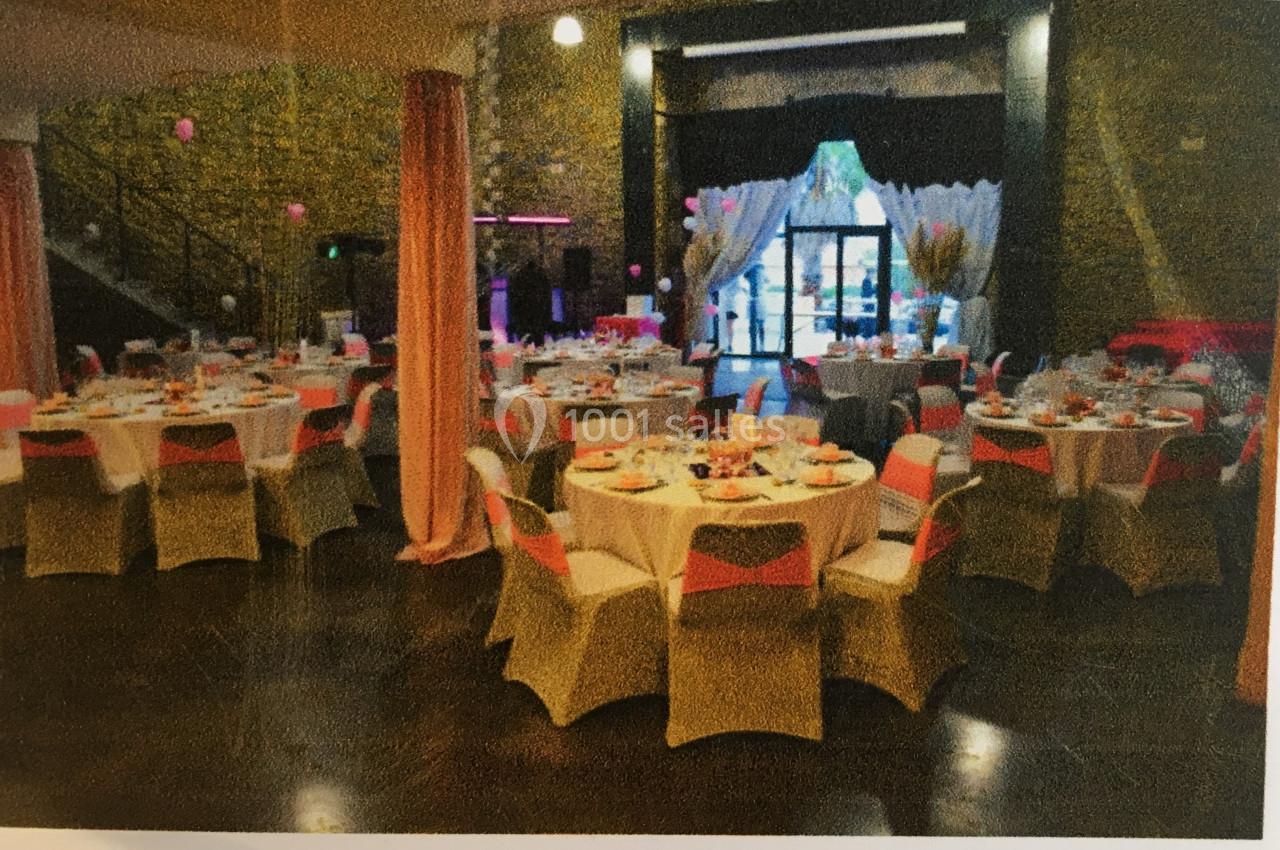 Salle de réception décorée avec des tables rondes dressées, nappes blanches et chaises ornées de rubans roses.