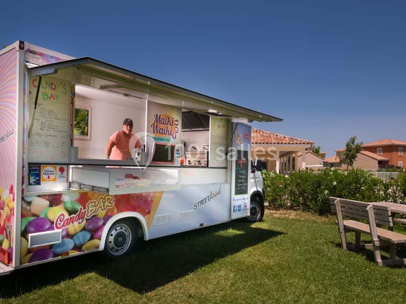 Un food truck coloré stationné sur une pelouse, avec un vendeur à l'intérieur et des maisons en arrière-plan.