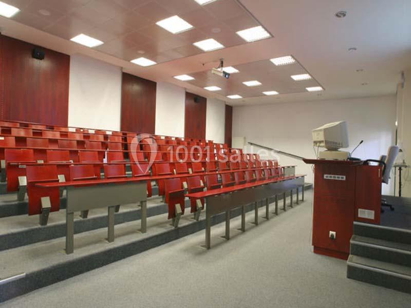 Salle de conférence vide avec rangées de sièges rouges, un pupitre et un projecteur au plafond.