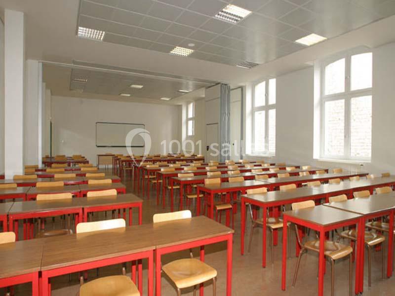 Salle de classe vide avec des tables en bois, des chaises et un tableau blanc, éclairée par des fenêtres et des néons.