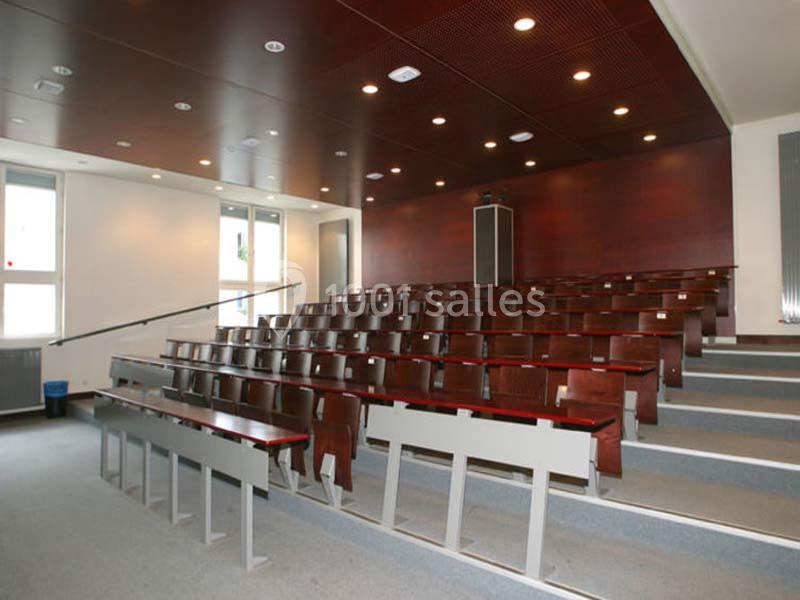 Salle de classe vide avec rangées de bancs en bois, murs clairs et plafond en bois sombre éclairé par des spots.