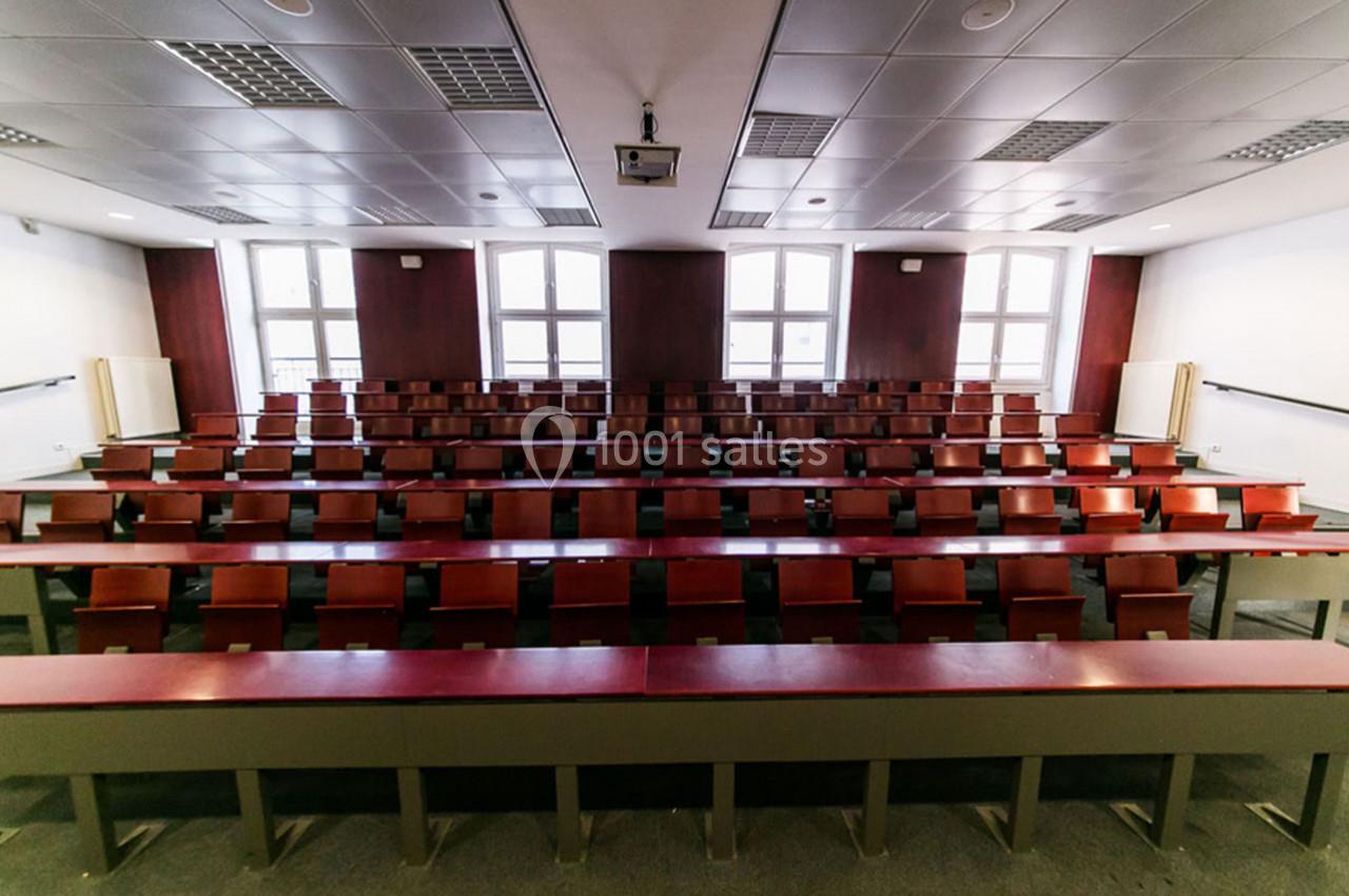 Salle de classe vide avec rangées de tables et chaises en bois, éclairée par des fenêtres et des lumières au plafond.