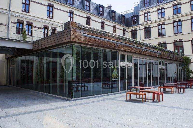 Cour intérieure avec bâtiment moderne en verre et bois, entouré d'édifices anciens, tables rouges disposées à l'extérieur.