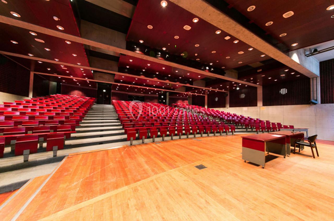 Salle de conférence vide avec rangées de sièges rouges, scène en bois et éclairage au plafond.
