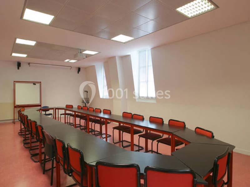 Salle de réunion avec une grande table en U entourée de chaises rouges, éclairée par des fenêtres et des plafonniers.