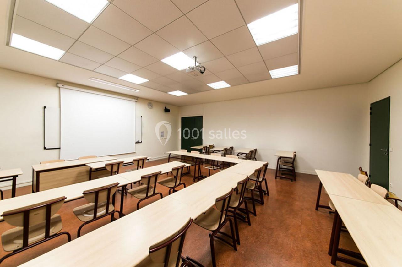 Salle de classe vide avec des rangées de tables et chaises, tableau blanc et projecteur au plafond.