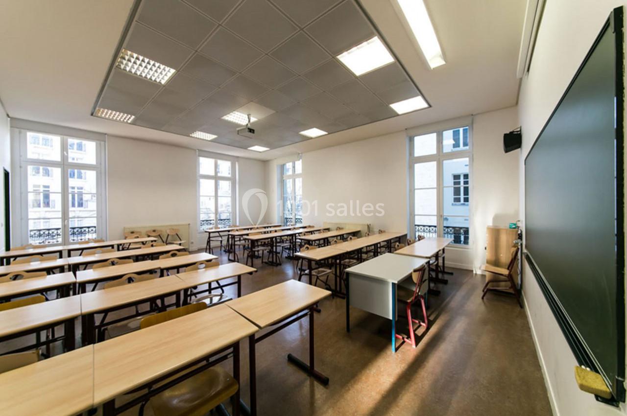Salle de classe lumineuse avec des tables en bois alignées, un tableau noir et de grandes fenêtres donnant sur l'extérieur.