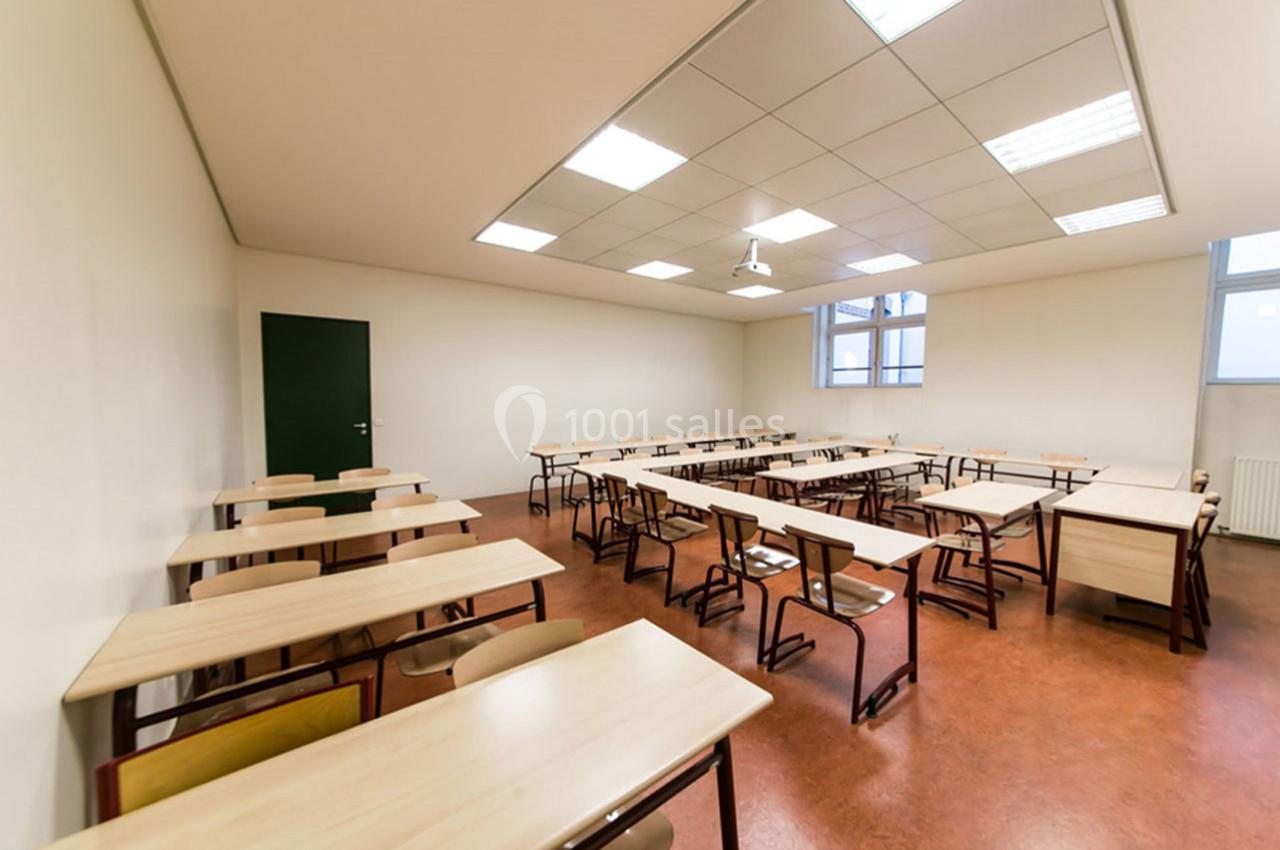 Salle de classe vide avec des bureaux en bois alignés, sol en lino rouge et plafond avec éclairage fluorescent.