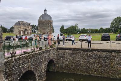 Façade en pierre d'un château bordé par un cours d'eau, avec un pont en arc et des arbres environnants.