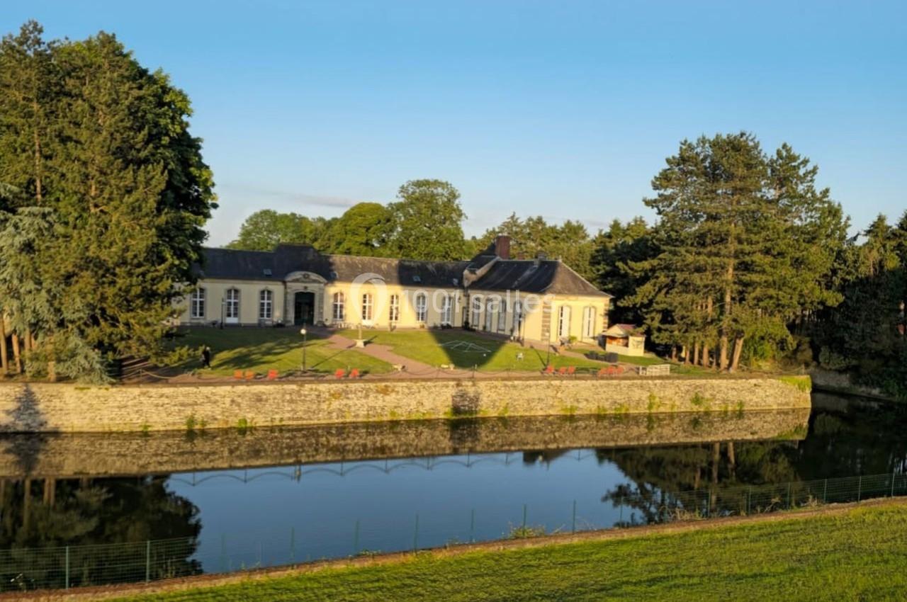 Bâtiment historique entouré d'arbres, avec un plan d'eau au premier plan reflétant la lumière du jour.